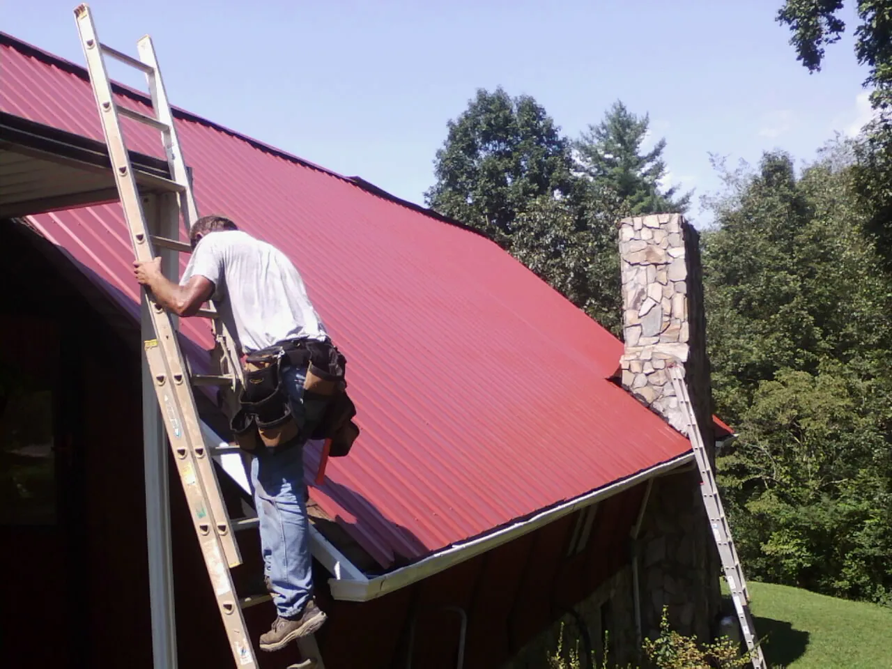 Roofer on a ladder beside a red metal roof and stone chimney — Soffit & Fascia Repair by Roofing Brothers in Ririe